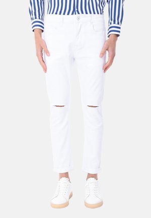 Persona che indossa jeans bianchi strappati risvoltati alle caviglie, scarpe da ginnastica bianche con suola in gomma e una camicia a righe bianca e blu.