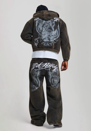 Homme portant une veste en denim foncé et un pantalon avec des motifs de crâne et d'ailes ainsi qu'un texte en écriture blanche au dos, debout devant un fond uni.
