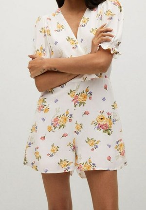 Nainen, joka pukeutunut valkoiseen kukkakuosiseen haalariin, jossa on keltaisia, vaaleanpunaisia ja violetteja kukkia, liehukkeet lyhyissä hihoissa ja V-aukko, kädet ristissä rinnalla.