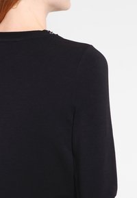 Černý top s dlouhým rukávem, hladká bavlněná textura, malé ozdobné knoflíky na zádech, kulatý výstřih a přiléhavý střih.