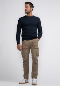 Marineblauwe gebreide trui, taupe cargobroek met zakken en witte sneakers met zwarte strepen. Model staat tegen een neutrale achtergrond.