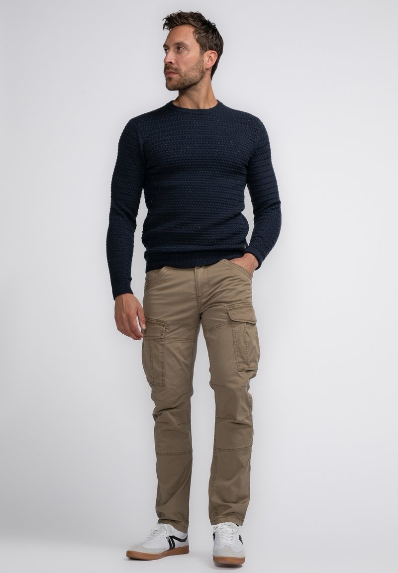 Marineblauwe gebreide trui, taupe cargobroek met zakken en witte sneakers met zwarte strepen. Model staat tegen een neutrale achtergrond.