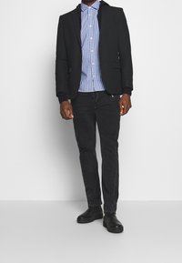 Zwarte blazer over een blauw-wit geruit shirt, gecombineerd met zwarte jeans en zwarte schoenen. Heeft een slim fit en een gladde, gestructureerde stof.