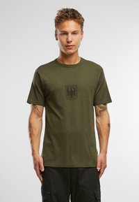 Olive zaļas krāsas kokvilnas T-krekls ar īsām piedurknēm, apaļu izgriezumu un melnu drukātu Bundeswehr logo priekšpusē. Vienkāršs dizains, nav redzamu rakstu.