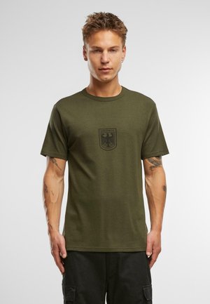 Camiseta de algodón verde oliva con mangas cortas, cuello redondo y un logo impreso de la Bundeswehr en negro en la parte delantera. Diseño simple, sin patrones visibles.