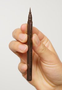 Maybelline Eyeliner-Stift mit einer feinen, abgeschrägten Spitze, braunem Gehäuse und schwarzem Schriftzug. Senkrecht in einer Hand gehalten vor einem hellen Hintergrund.
