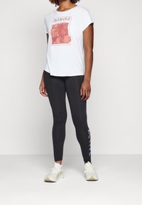 Biała koszulka z kwiatowym nadrukiem i napisem "Bądź świadomy", zestawiona z czarnymi legginsami z wielokolorowym napisem "ACTIVE" po boku.