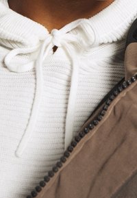 Biała teksturowana bluza z kapturem z zapięciem na wiązanie przy szyi, noszona pod brązową kurtką na zamek z okrągłymi czarnymi elementami metalowymi.