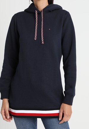 Personne portant un sweat à capuche bleu marine avec des cordons à motifs et des rayures rouges, blanches et noires à l'ourlet, associé à un jean bleu.