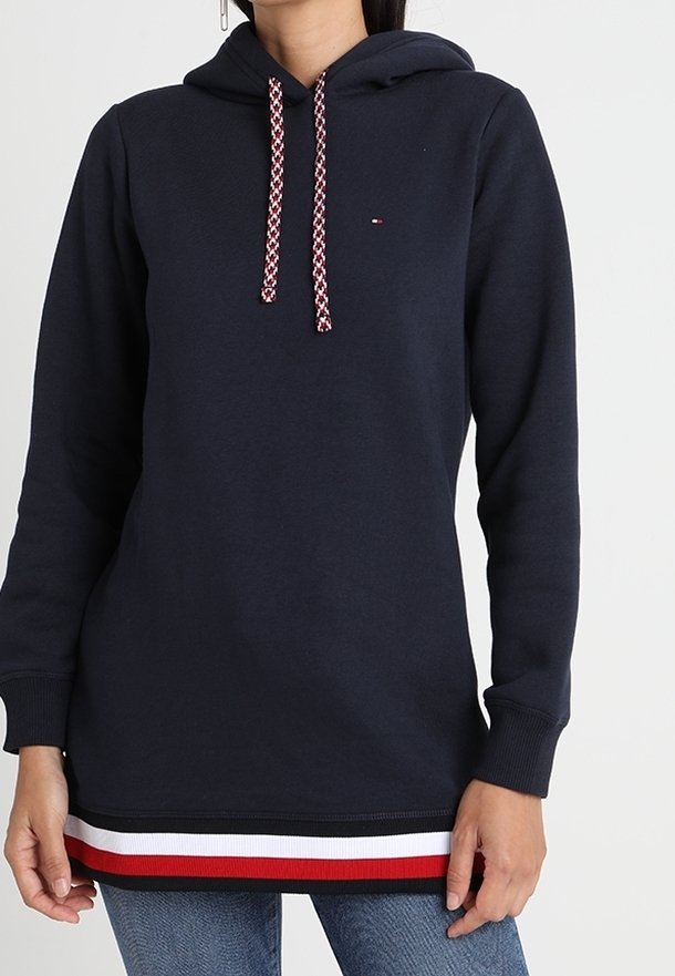Personne portant un sweat à capuche bleu marine avec des cordons à motifs et des rayures rouges, blanches et noires à l'ourlet, associé à un jean bleu.