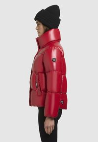 Rode gewatteerde jas met een hoge kraag, glanzende textuur, zijzakken en een logopatch. Draag met een zwarte gebreide beanie.