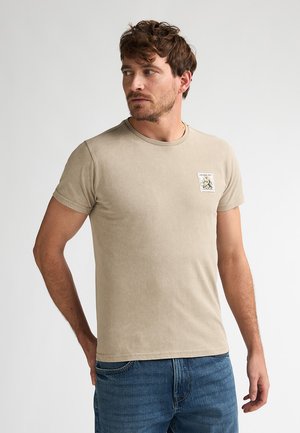 T-shirt beige à manches courtes avec un col rond et un petit patch graphique sur le côté gauche de la poitrine. Texture douce, porté avec un jean bleu.