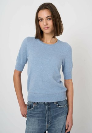 Jonge vrouw met schouderlang bruin haar, gekleed in een lichtblauwe trui met korte mouwen en een blauwe spijkerbroek, tegen een effen achtergrond.