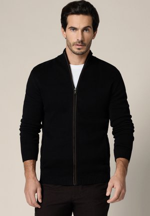 Mężczyzna o ciemnych włosach, ubrany w czarny sweter na zamek błyskawiczny na białą koszulę, stojący na jednolitym beżowym tle.