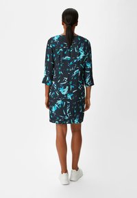 Korte mouw jurk van marineblauwe stof met turkooise bloemenpatroon. Heeft een rechte snit en valt boven de knie, gecombineerd met witte sneakers.