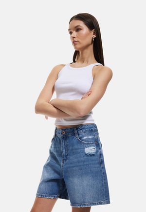 Denim-Shorts mit einem ausgebleichten blauen Waschung, fransigem Saum und abgenutzten Details am rechten Oberschenkel, kombiniert mit einem enganliegenden weißen Tanktop.
