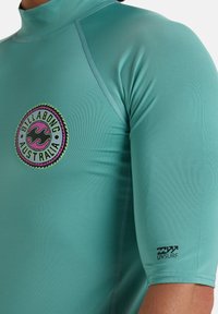 Rash guard en color azul verdoso con un cuello alto, mangas cortas y un logo circular de Billabong en rosa y verde. Presenta costuras planas para mayor comodidad.