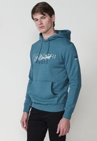 Felpa con cappuccio color teal e tasca a marsupio. Presenta grafiche testuali in bianco e oliva. Progettata con polsini e bordo a coste.