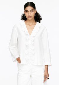 White cotton blouse featuring a deep V-neck, ruffled front placket, and long sleeves. Smooth texture and loose fit for comfort.