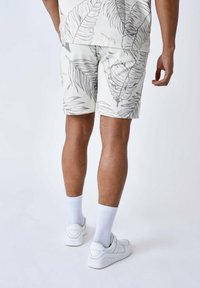 Shorts en un fondo claro con un patrón de hojas de palma grises. Hechos de tela, presentan un corte relajado y se estilizan con zapatillas deportivas y calcetines blancos.