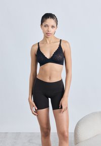Μαύρο δαντελένιο σουτιέν με κυματιστές άκρες και ρυθμιζόμενους ιμάντες, σε συνδυασμό με μαύρο εφαρμοστό σορτς, πάνω σε γκρι φόντο.