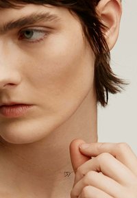 Collar de oro con un colgante de doble corazón, cadena delicada, llevado sobre piel desnuda, mostrado cerca de una mano tocando el colgante. Primer plano del diseño.