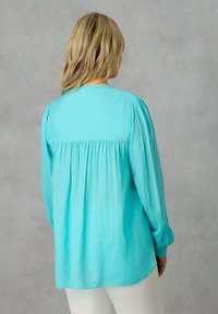 Blusa di un leggero turchese con maniche lunghe, design a pieghe sul retro e una stoffa morbida e testurizzata. Collo rotondo liscio e vestibilità leggermente ampia.