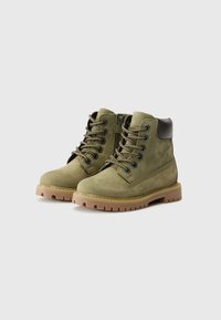 Bottines en suède vert avec des accents noirs, devant à lacets et semelles en caoutchouc texturées. Présente des détails cousus et un col rembourré.