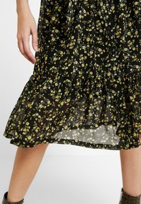 Robe noire à motifs floraux avec des dessins jaunes et verts, couches transparentes, ourlet froncé et détails de boutons près de la taille. Tissu texturé.