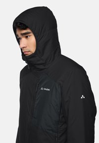 Schwarze isolierte Jacke mit Kapuze, mit einem Reißverschluss auf der Vorderseite und kontrastierendem dunklem Stoff auf der Brust. Logo am Oberarm. Weiche Textur.