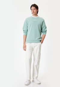 Sudadera verde menta con "CONFIDENT" en relieve en el pecho, combinada con pantalones cargo blancos y zapatillas blancas.
