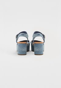 Blå denim plattformshelar med öppen baksida, vadderade remmar och en bred blockklack. Texturerad yta med en naturlig brun sula.