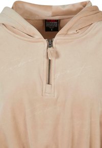 Bežová mikina na zips vyrobená z jemného materiálu, s predným vreckom, kapucňou a diskrétnymi bielymi textovými vzormi po celom povrchu.