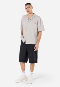 Cămașă cu mânecă scurtă, de culoare gri deschis, cu material texturat, două buzunare frontale și un logo contrastant al brandului. Purtați cu pantaloni scurți negri și adidași albi.