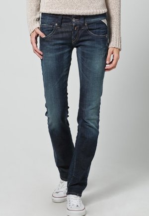 Jean en denim bleu foncé avec une coupe ajustée, légères décolorations, cinq poches et petits éléments en métal. Associés à des baskets blanches.