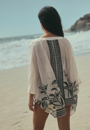 Femeie cu păr lung și închis la culoare, purtând un costum alb de plajă cu model, stând pe o țărm nisipos, cu fața spre ocean, cu stânci în fundal.