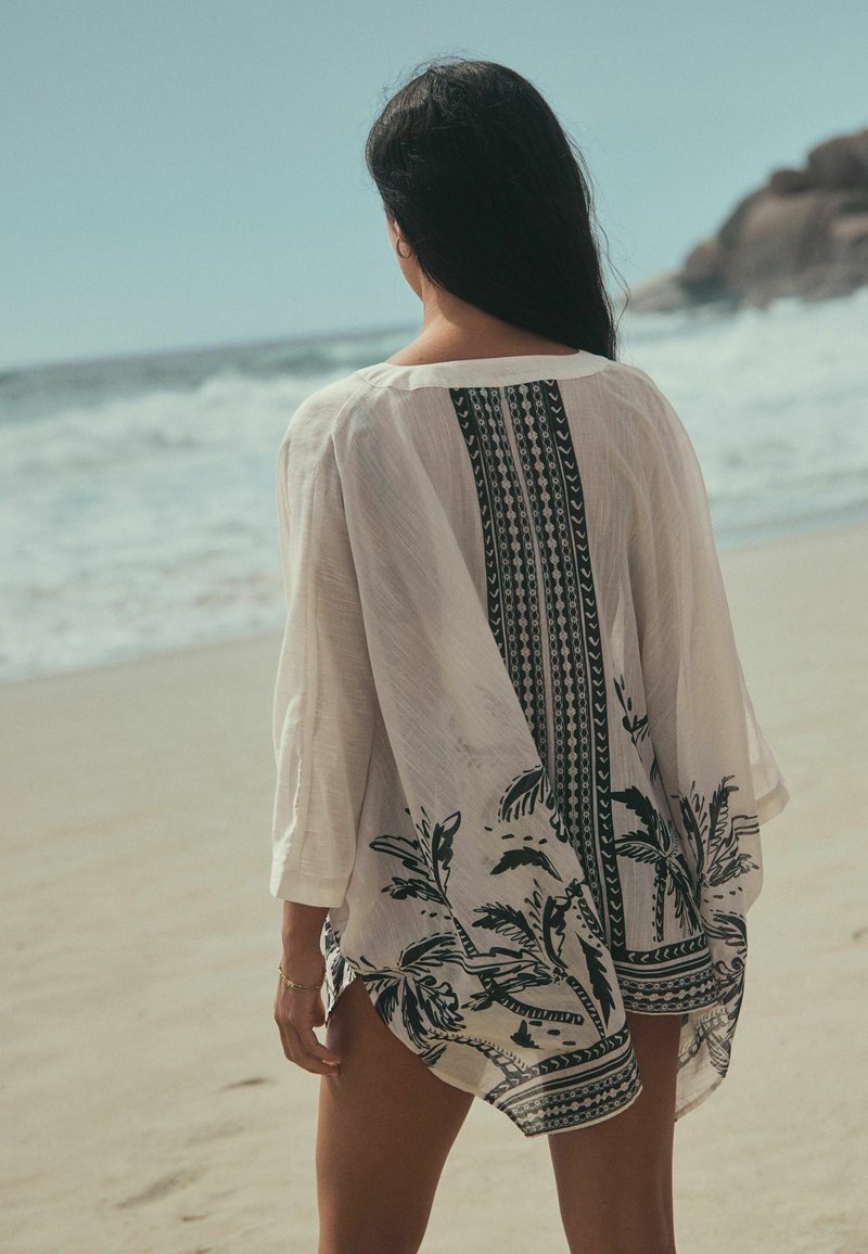 Femme aux longs cheveux foncés portant un pareo de plage blanc à motifs, debout sur une plage sableuse face à l'océan avec des rochers en arrière-plan.