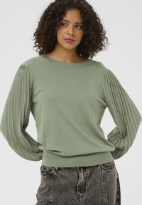 Pulover tricotat verde deschis, cu mâneci cu pliuri și tivuri drepte. Guler rotund și tiv croit strâns, cu detaliu texturat. Modelul este asortat cu blugi de culoare închisă.