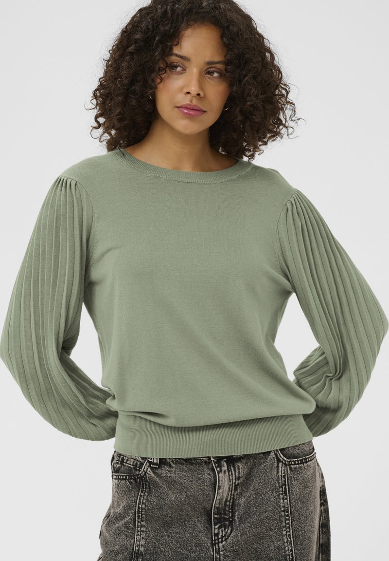 Pulover tricotat verde deschis, cu mâneci cu pliuri și tivuri drepte. Guler rotund și tiv croit strâns, cu detaliu texturat. Modelul este asortat cu blugi de culoare închisă.