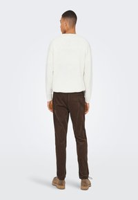 Weißer Pullover mit strukturiertem Muster, kombiniert mit braunen Cordhosen. Modell steht mit dem Rücken zur Kamera, trägt hellbraune Schuhe, neutraler Hintergrund.