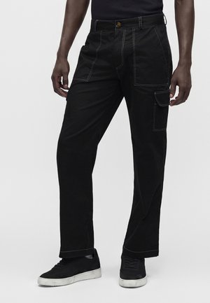 Hombre con pantalones cargo negros con costuras blancas y zapatillas negras con suelas blancas, de pie frente a un fondo liso.