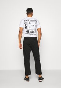 Bílý tričko s černým grafickým textem a logem na zádech, spárované s černými kalhotami a černými teniskami s bílými podrážkami.