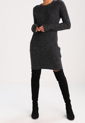 Mujer con vestido de punto acanalado gris oscuro y botas negras por encima de la rodilla, de pie con las manos entrelazadas al frente.