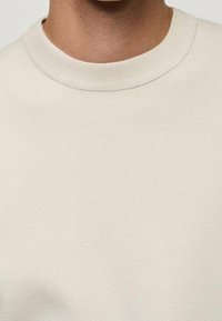 Gros plan sur une personne portant un pull col rond de couleur crème, montrant l'encolure et la partie supérieure de la poitrine.