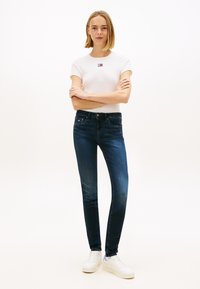 Jeans justos azul escuro combinados com uma t-shirt branca de mangas curtas com um pequeno logótipo vermelho e azul, usados com sapatilhas brancas.