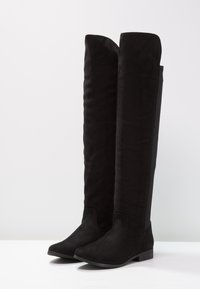 Botas negras por encima de la rodilla hechas de suave ante con suela plana, con una textura lisa y diseño de punta redondeada.