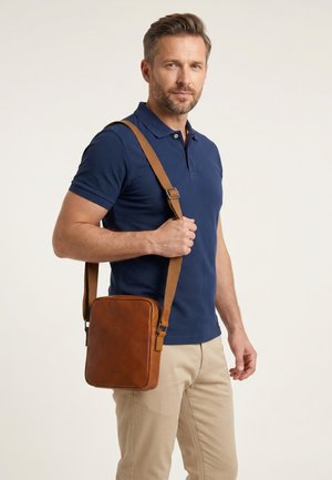 Man in een marineblauw polo en beige broek draagt een bruine leren crossbodytas met verstelbare band over de schouder.