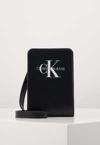 Černá obdélníková crossbody taška Calvin Klein Jeans s bílým logem a tenkým popruhem, stojící na bílém povrchu.