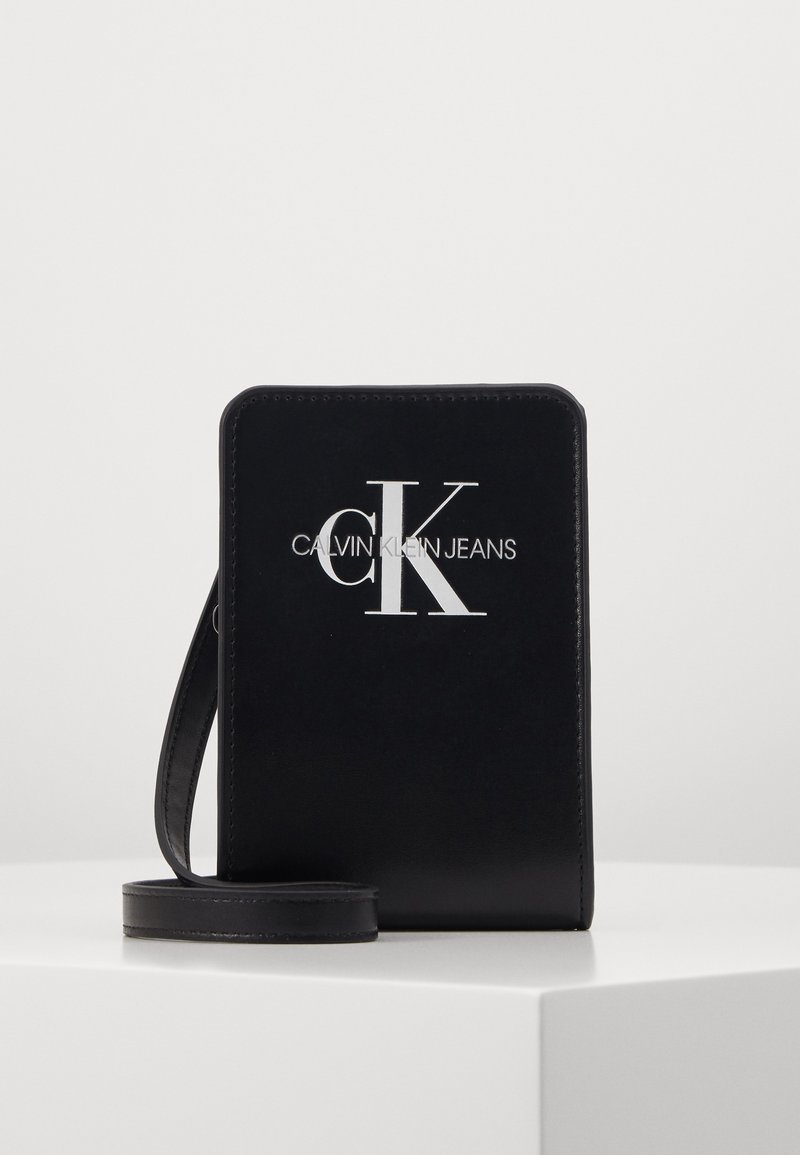 Černá obdélníková crossbody taška Calvin Klein Jeans s bílým logem a tenkým popruhem, stojící na bílém povrchu.