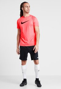 Herren Nike Sportoutfit bestehend aus einem leuchtend pinkfarbenen strukturierten T-Shirt mit kurzen Ärmeln und schwarzem Logo, kombiniert mit schwarzen Shorts und weißen Kniestrümpfen.